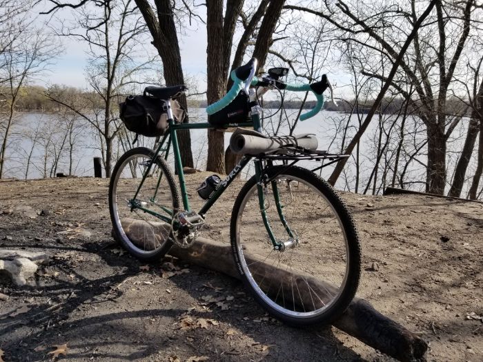 Angled front side view of a green Surly Rat Pack bike standing upright on a dirt clearing with trees and lake behind