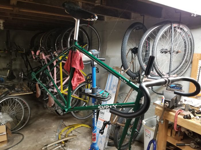 Ride side view of a green Surly Rack Rat bike frame with various parts on a repair stand in a basement with bikes and parts