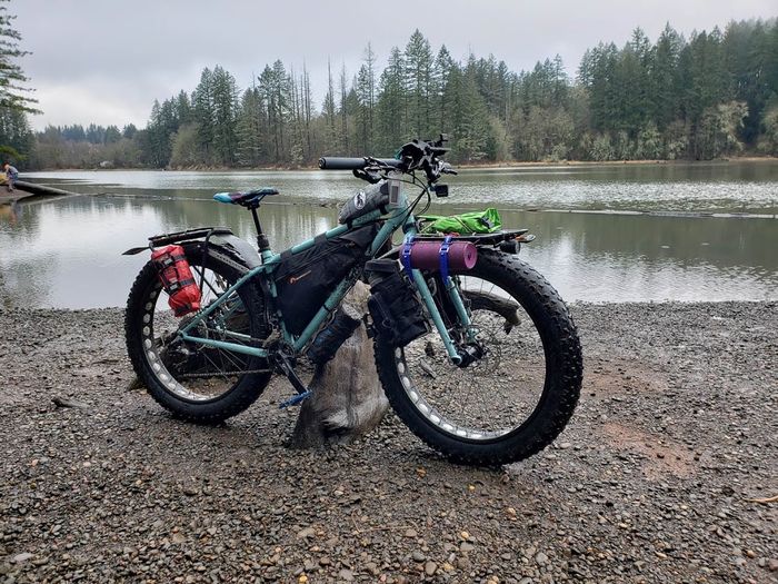 Lefty side view of a Surly Ice Cream Truck bike, mint, on a gravel shore of a lake surrounded with pine trees