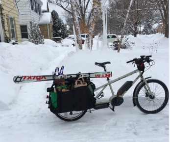 Right profile view of a tan Surly Big Easy bike with rear saddle and skis in the snow with trees and house in background