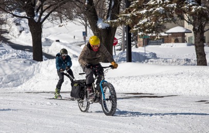 Cyclist riding a fat bike towing a skier down a snow covered street in a neighborhood with trees