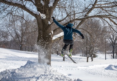 A skier in a hooded blue coat, flies through the air near a tree, after launching from a jump in a snow covered field