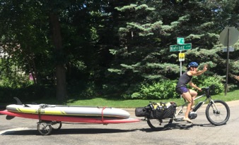 Cyclist riding a tan Surly Big Easy bike with rear saddle bags pulling two paddle boards on a trailer down a street