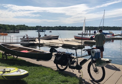 Right side view of a tan Surly Big Easy bike with trailer carrying a red canoe on a dock beside a lake with a few boats