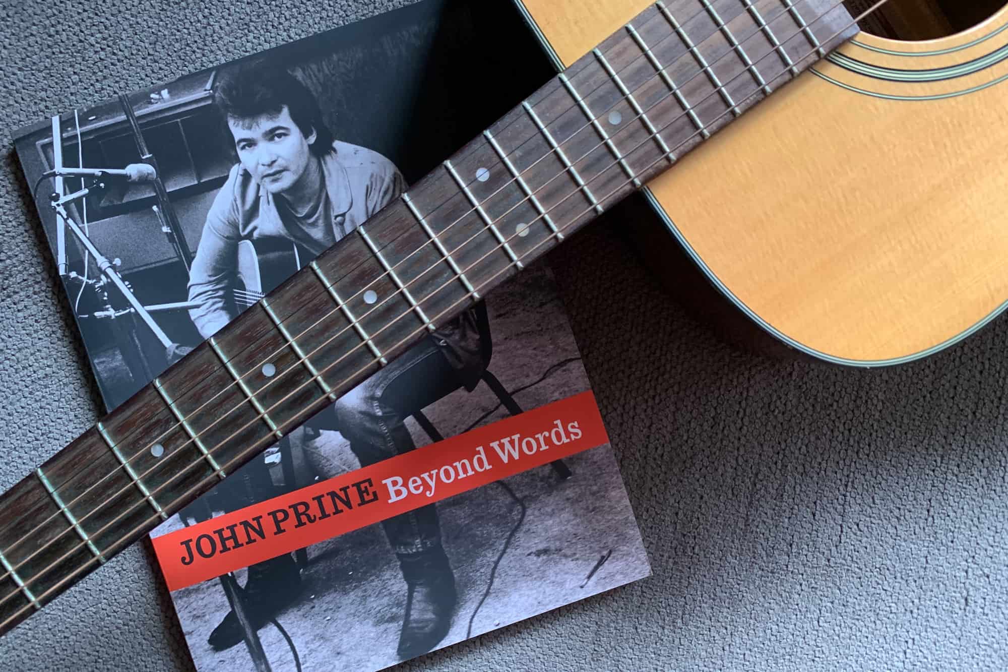 Portrait of John Prine book Beyond Words lying on short gray carpet with the neck of an acoustic guitar laying across