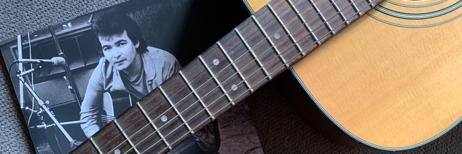 Portrait of John Prine book Beyond Words lying on short gray carpet with the neck of an acoustic guitar laying across