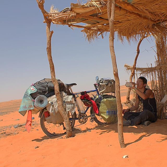 Cyclist sitting on red sand drinking water under a grass and stick canopy next to a blue Surly bike loaded with gear