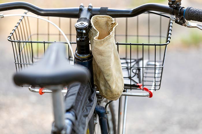 Rear view of bike showing homemade leather handlebar feed bag, rack/basket, handlebar with bell, brake levers and lock-on grips with blue collars and saddle