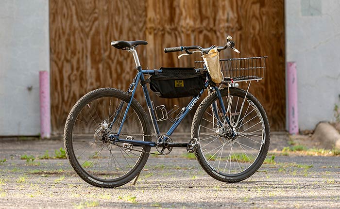 Surly Straggle Rat rear three-quarter view of single speed bike with two water bottle cages, frame bag, handlebar feed bag, and front rack with basked in front of boarded up building