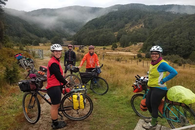 Monica with three other cyclists on gravel road with loaded bikes, mountains in background