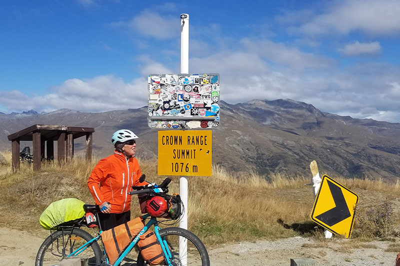 Monica with light blue fully loaded Surly Bridge Club a Crown Range Summit sign with mountains in background