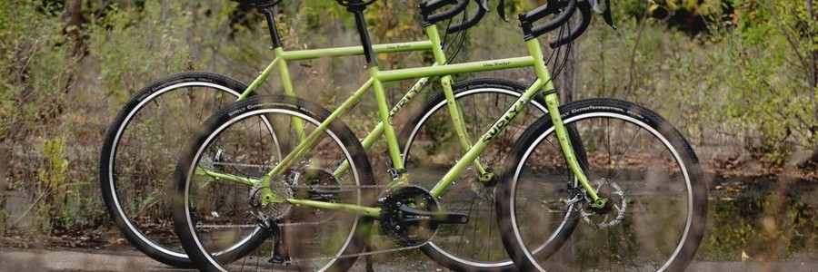 Right side view of 2, pea lime soup colored, Surly Disc Trucker bikes, side by side, on watery ground with brush behind