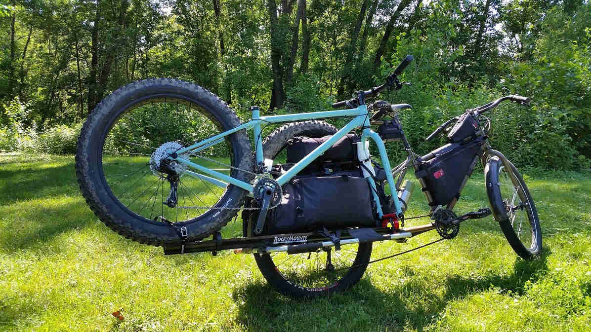 Rear, right side view of a Surly Big Dummy bike hauling a fat bike, parked in a grass field in front of trees