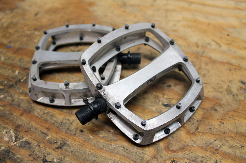 Silver, studded, platform bike pedals, with one resting on the other, laying on a wood workbench