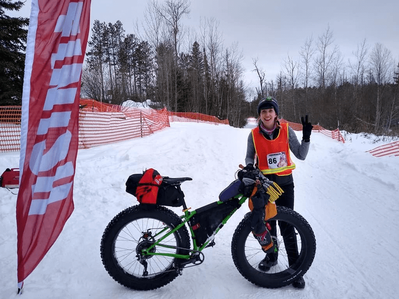 Cyclist smiles giving a peace sign while standing with  their fat bike, green, at a finish line of a snowy bike trail