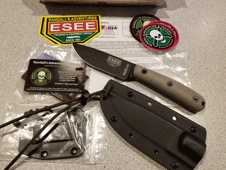 Black blade ESEE knife lying next to black sheath with Randall's Adventures & Training shirt patches and business card 