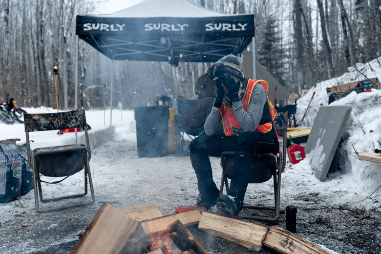 Person wearing mittens and winter gear cover their face while sitting at a chair at a snowy site in the woods