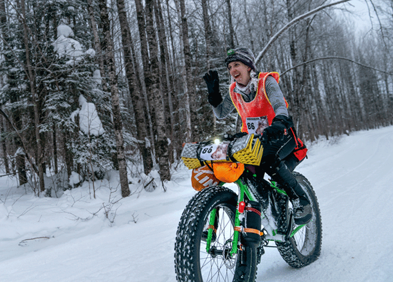 Angled left side view of a winter cyclist  raising their right hand riding down a snow covered hill trail in the woods