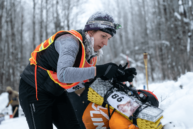 Amy dressed in winter clothing bends over a pack of biking gear on a snowy day in the woods