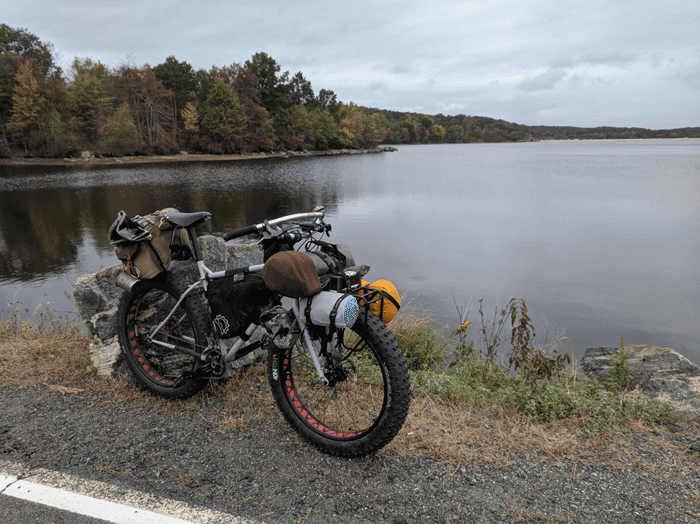 Right profile of a white Surly Pugsley fat bike loaded with gear on the shoulder of a paved road on the edge of a lake