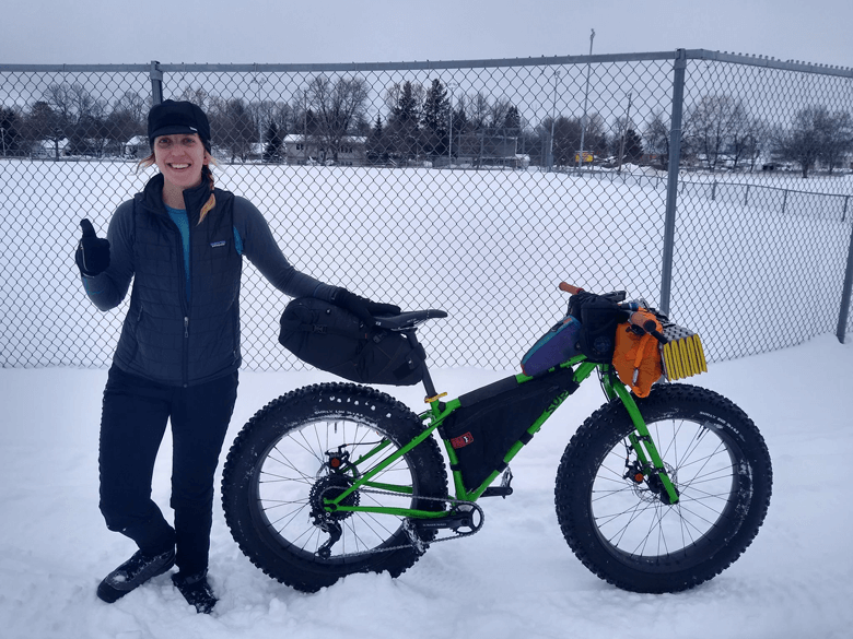 A cyclist poses at the back of a Surly fat bike, green, on a cloudy day in a snowy field with a chain link fence behind