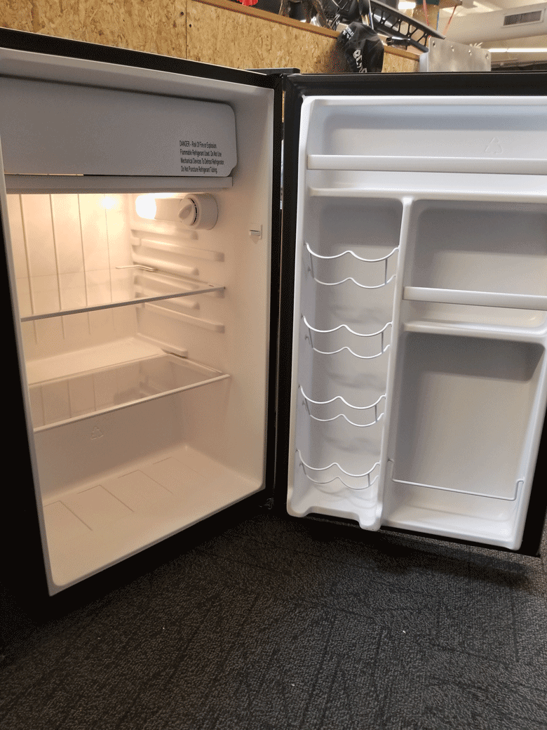 Angled view from the left side of a empty black refrigerator with fully open in front of an OSB wall